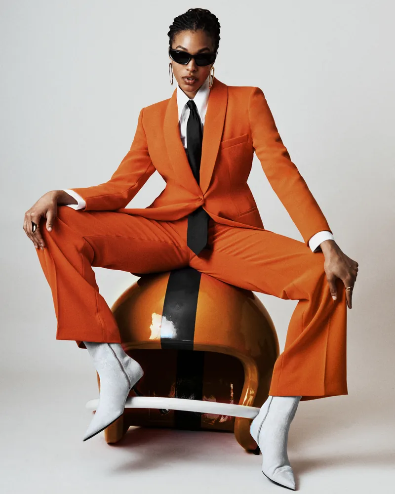 A model in orange tailoring and white boots seated on a sculptural chair.