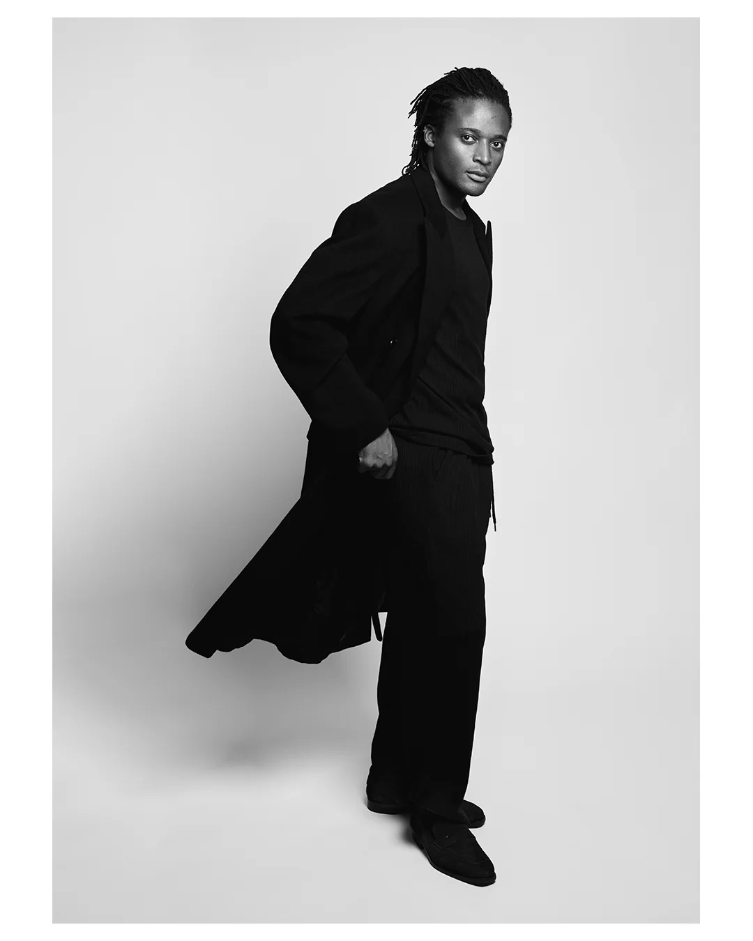 Black-and-white full-length portrait of Kudzai King in a black coat.