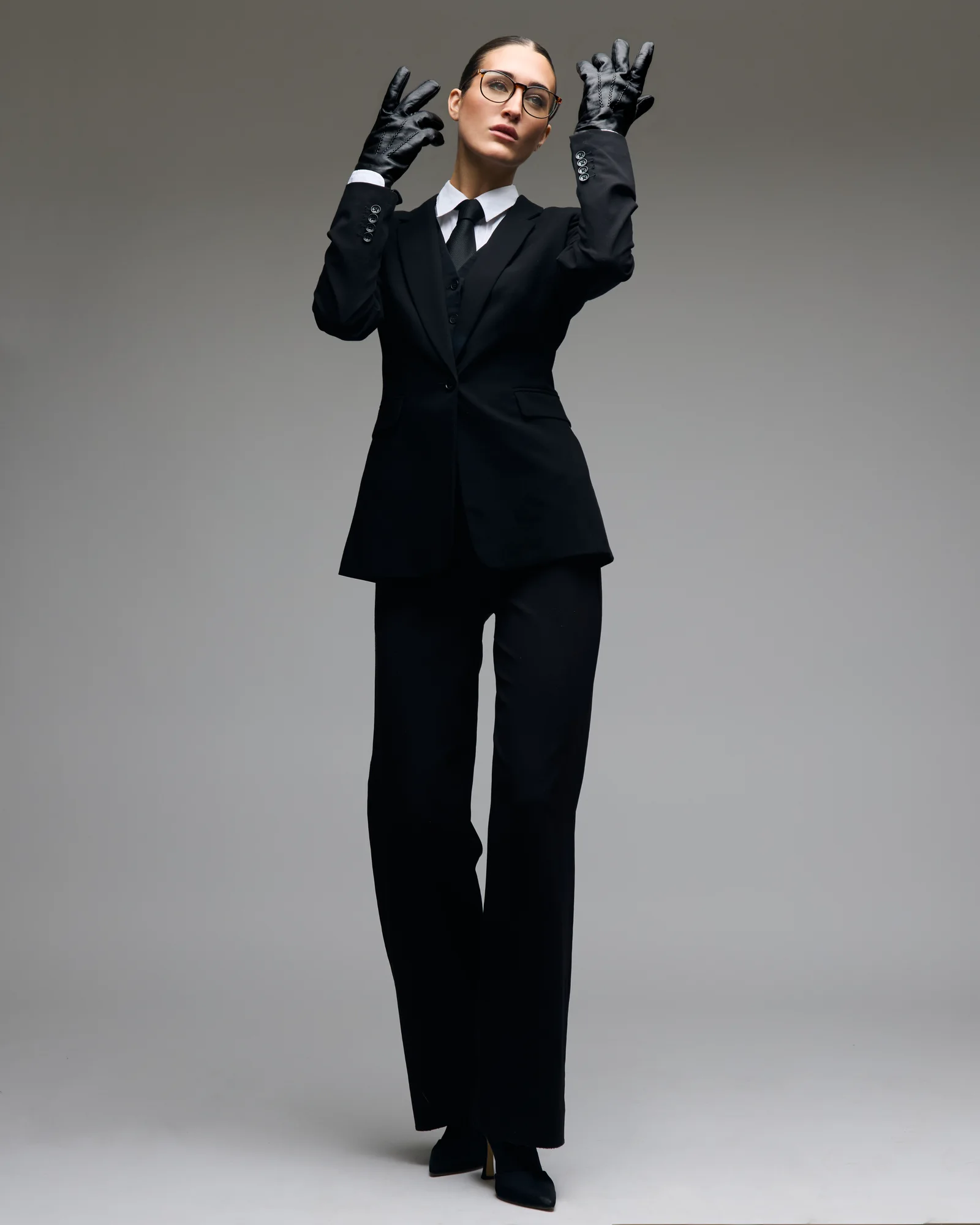 A studio portrait in black tailoring, glasses and gloves.