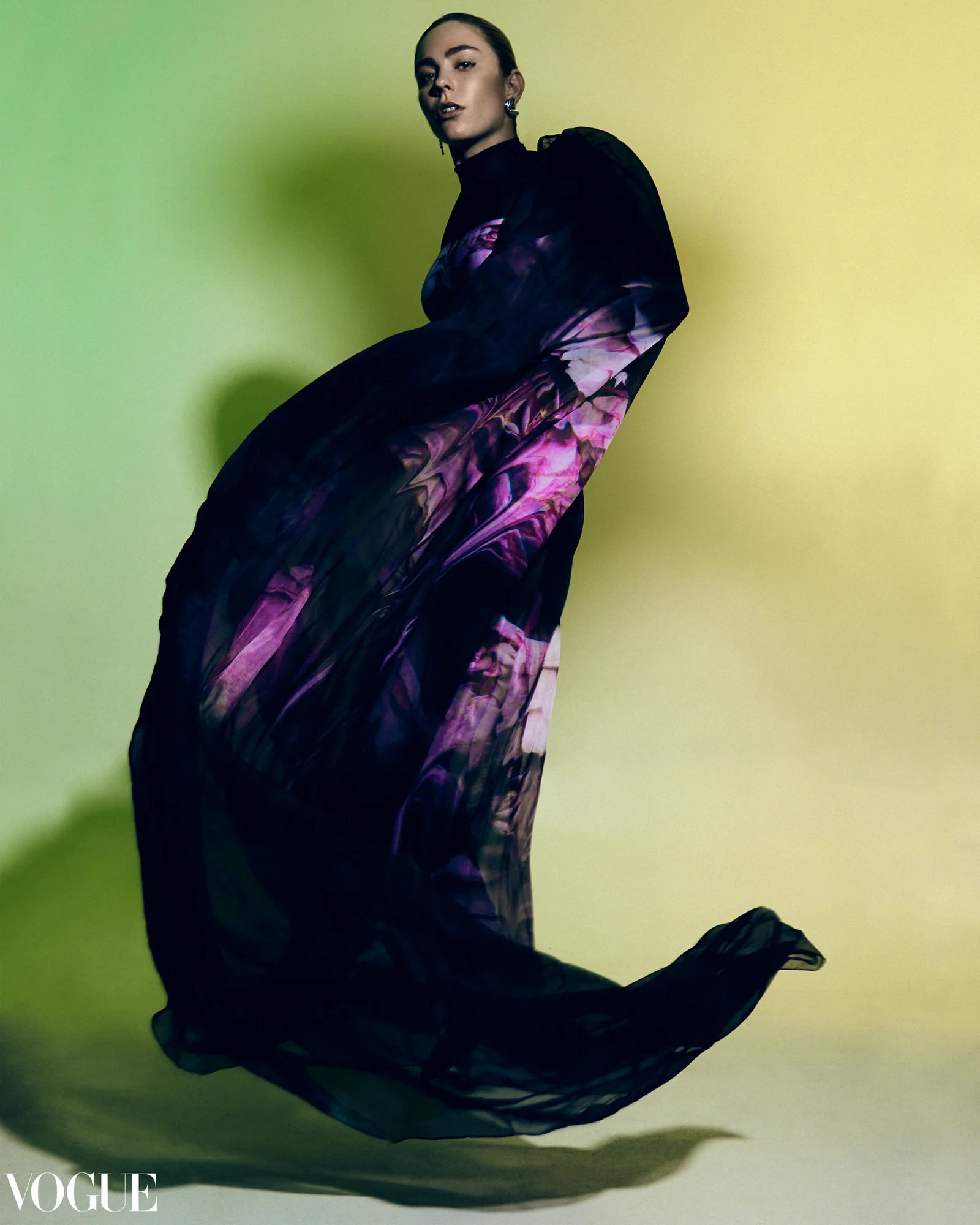 A model in a purple patterned dress with arcing fabric against green; the supplied image includes a Vogue wordmark.
