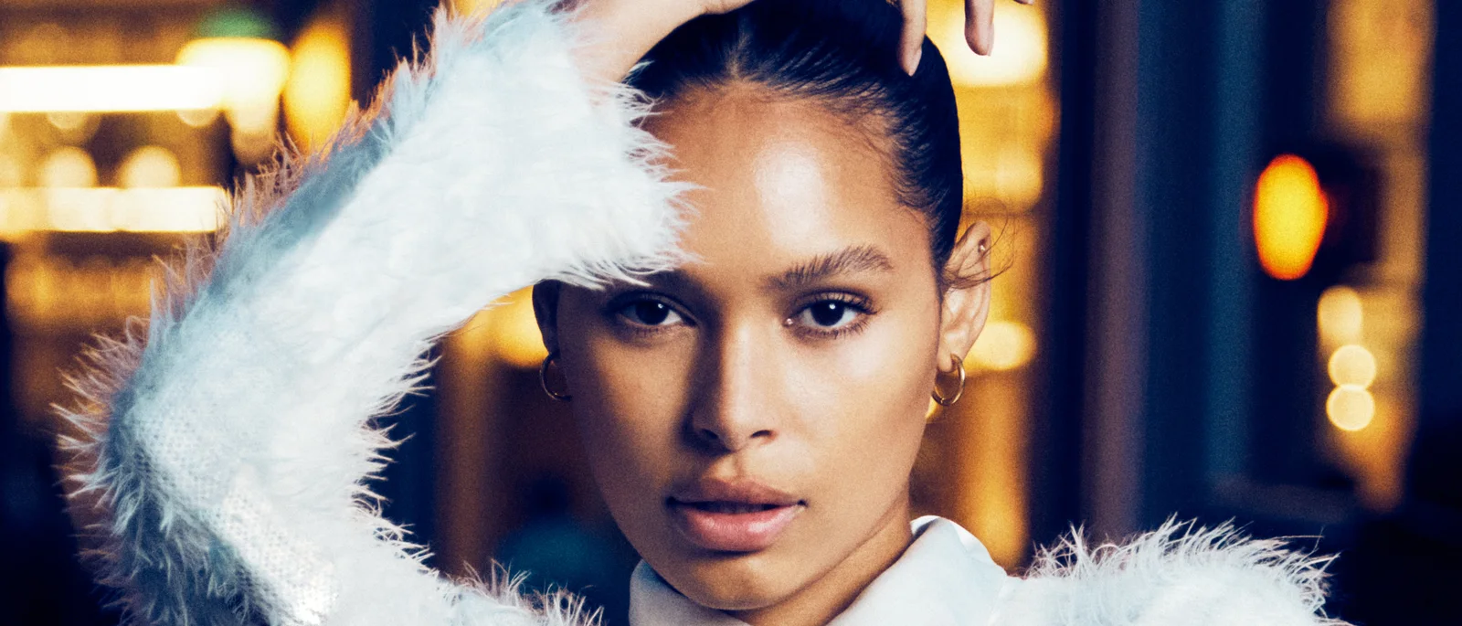 A close portrait framed by a white feather sleeve with warm city light behind.