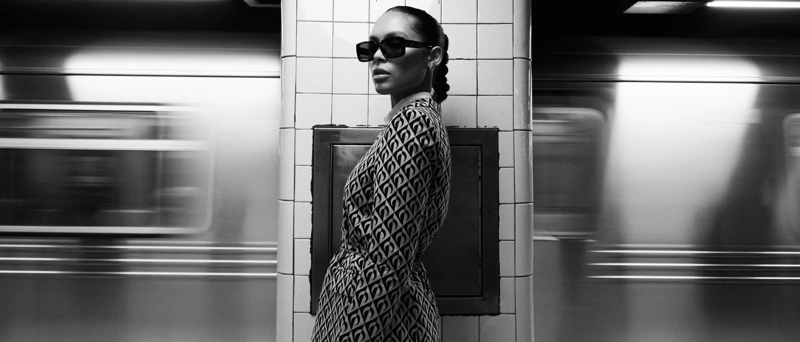 A model in patterned tailoring standing between motion-blurred trains in a black-and-white subway photograph.