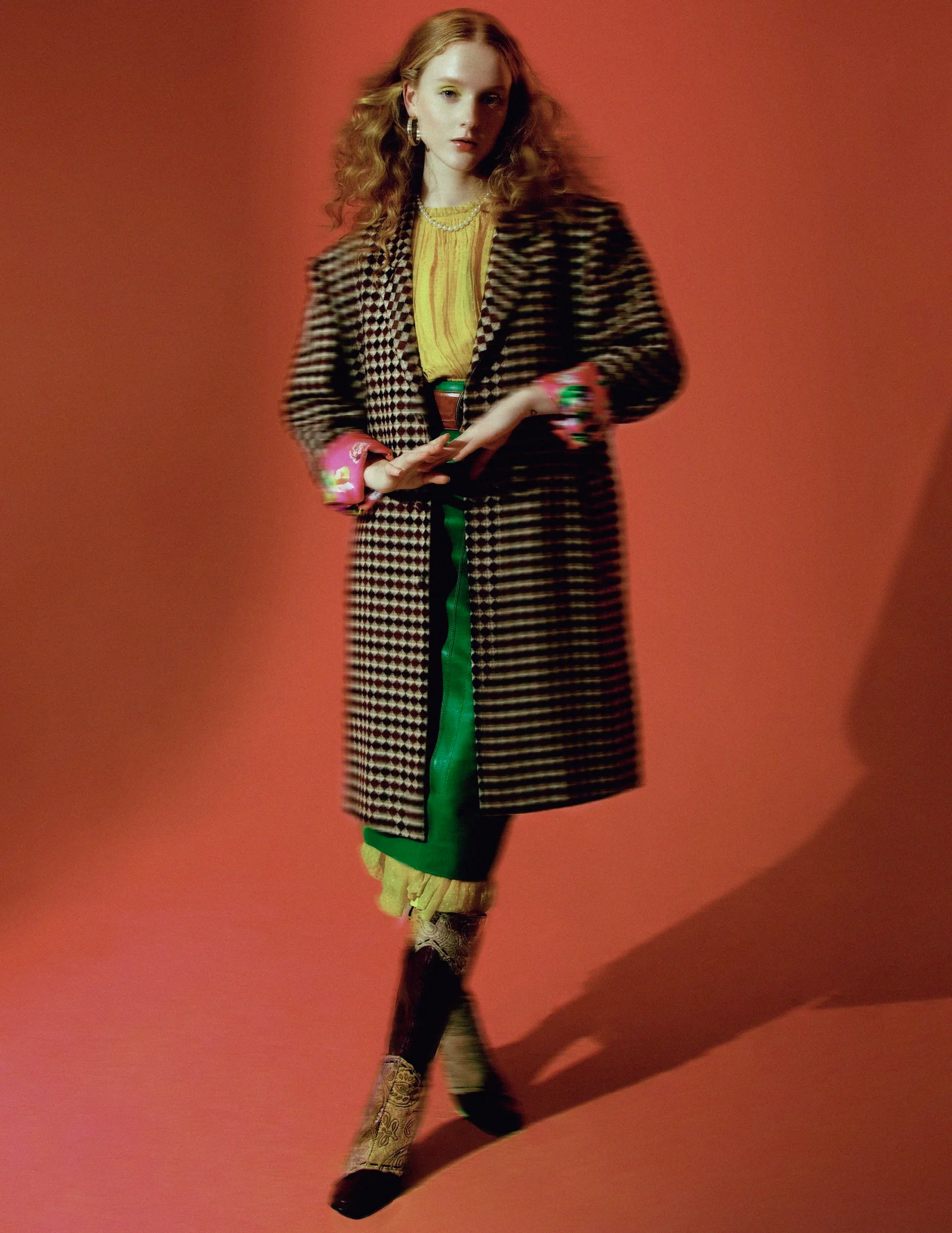 A model in a patterned coat and yellow top against an orange studio background.