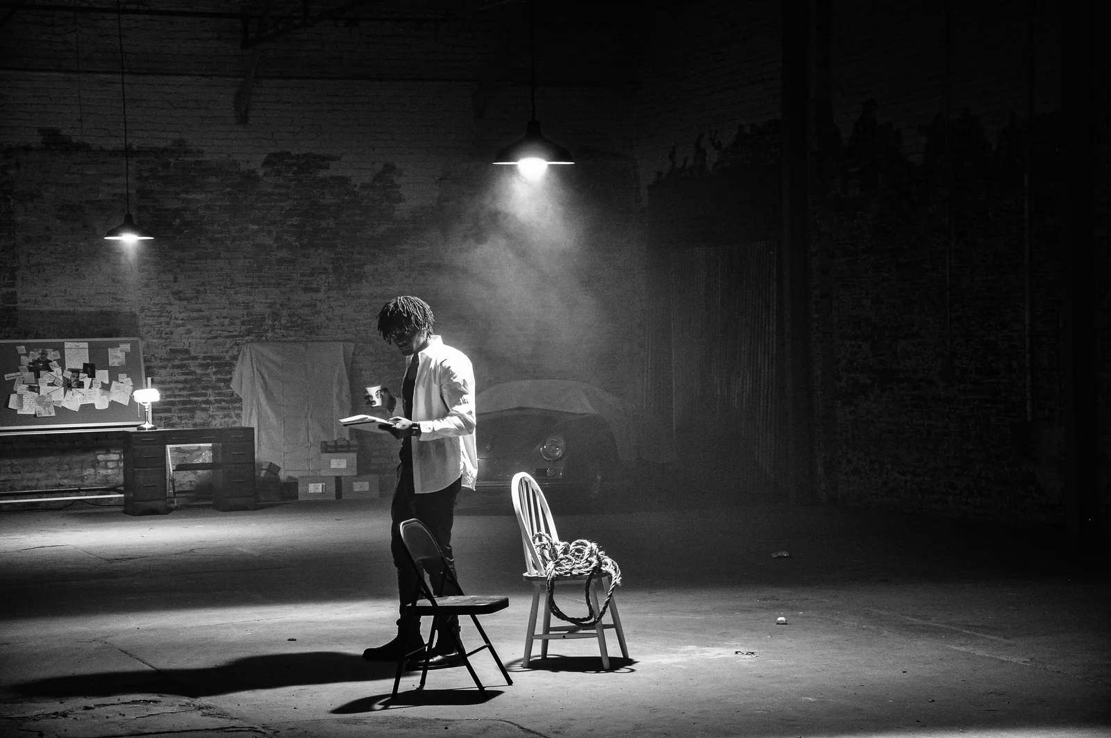 A wide black-and-white warehouse set with a working figure, chairs and overhead light.
